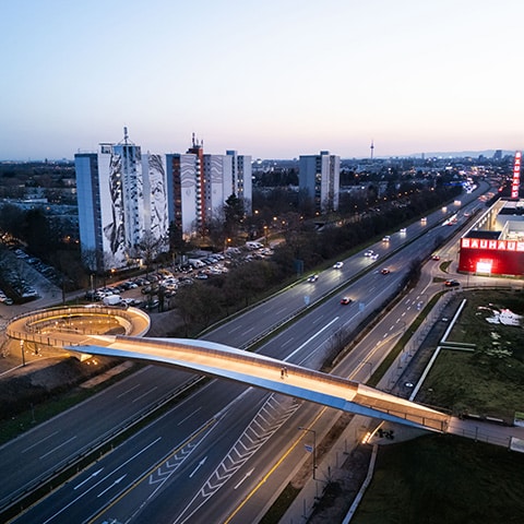 KI-basierte Bildbeschreibung | Eine geschwungene Fußgängerbrücke führt in der Abenddämmerung über eine mehrspurige Straße in der Nähe hoher Wohnblocks und eines hell erleuchteten Bauhaus-Geschäfts.