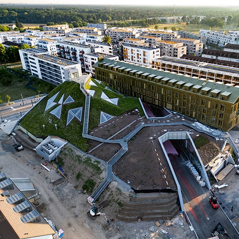 KI-basierte Bildbeschreibung | Luftaufnahme eines modernen Gebäudes mit einem grünen, kantigen Dach, geometrischen Oberlichtern und umliegenden städtischen Wohngebäuden.