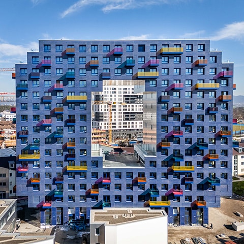 KI-basierte Bildbeschreibung | Ein quadratisches, blaues Wohnhaus mit einer großen Aussparung in der Mitte, das sich durch bunte Balkone auszeichnet und auf eine Baustelle sowie die Stadtlandschaft im Hintergrund blickt.