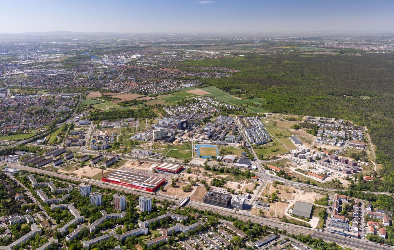 KI-basierte Bildbeschreibung | Luftaufnahme eines Vorortgebiets mit Wohngebäuden, Grünflächen und Baustellen, umgeben von Wäldern und einer in der Ferne liegenden Stadtlandschaft unter klarem Himmel.