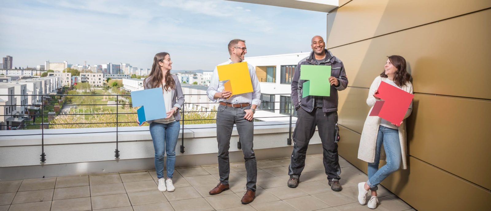 KI-basierte Bildbeschreibung | Vier Personen stehen auf einem Balkon, halten große, farbige Quadrate in der Hand und lächeln sich gegenseitig an, während im Hintergrund eine Stadtlandschaft zu sehen ist.