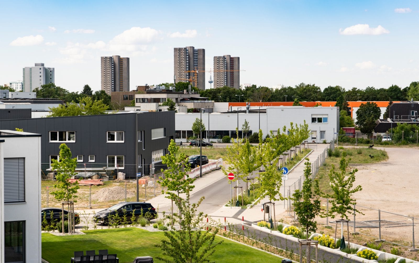KI-basierte Bildbeschreibung | Modernes Wohnviertel mit niedrigen Gebäuden, einer schmalen Straße, jungen Bäumen und Hochhauswohnungen im Hintergrund unter einem teilweise bewölkten Himmel.