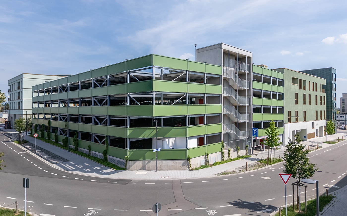 KI-basierte Bildbeschreibung | Ein mehrstöckiges, begrüntes Parkhaus mit Außentreppen, das an einer Straßenecke liegt, umgeben von Straßen und etwas Grün.
