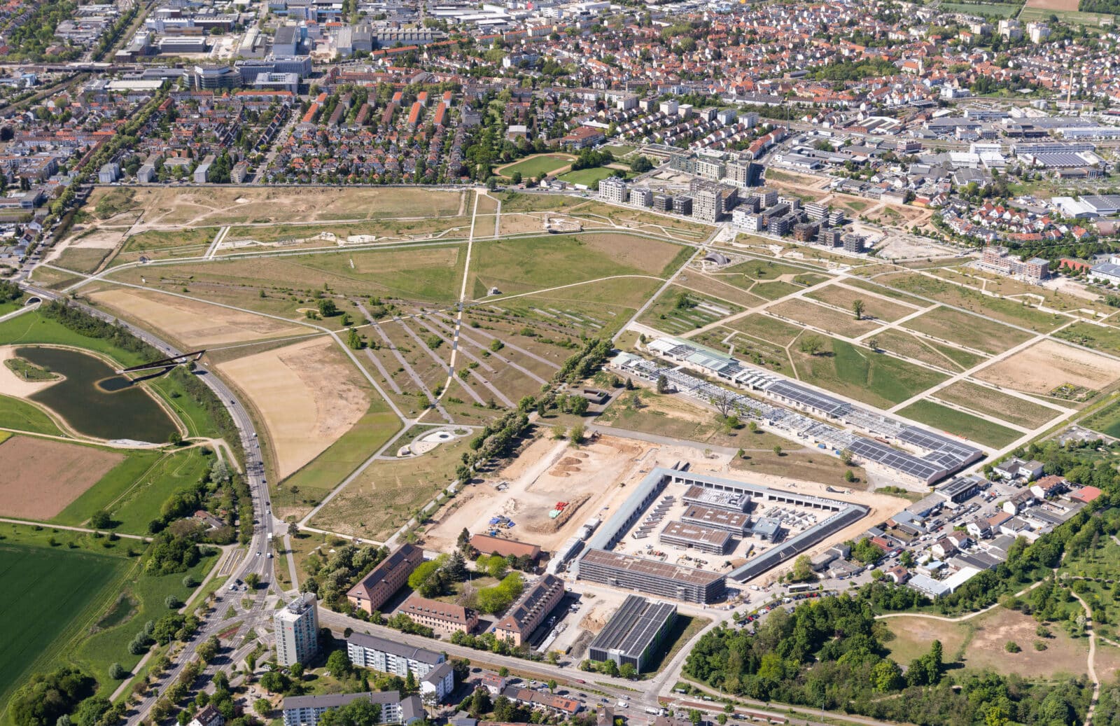 KI-basierte Bildbeschreibung | Luftaufnahme eines Stadtgebiets mit Wohngebäuden, offenen Feldern, Baustellen, Straßen und Grünflächen.